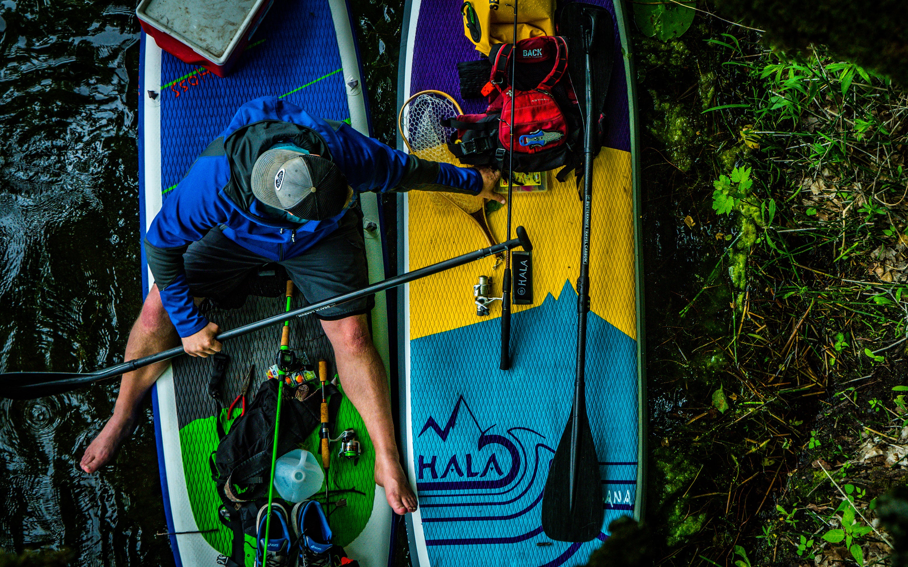 A high-angle shot of two Hala stand-up paddleboards (SUPs) rigged for SUP fishing and overnight touring, resting side-by-side on the edge of a river.