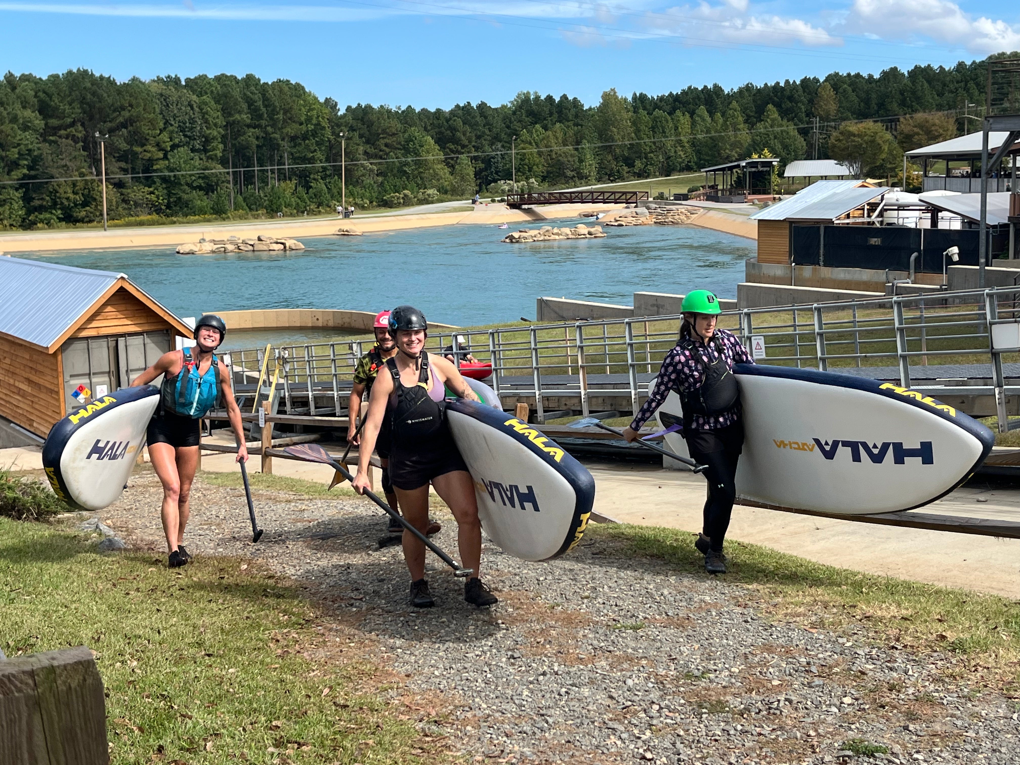 People carrying Hala paddleboards at the US National Whitewater Center with trees in the background.